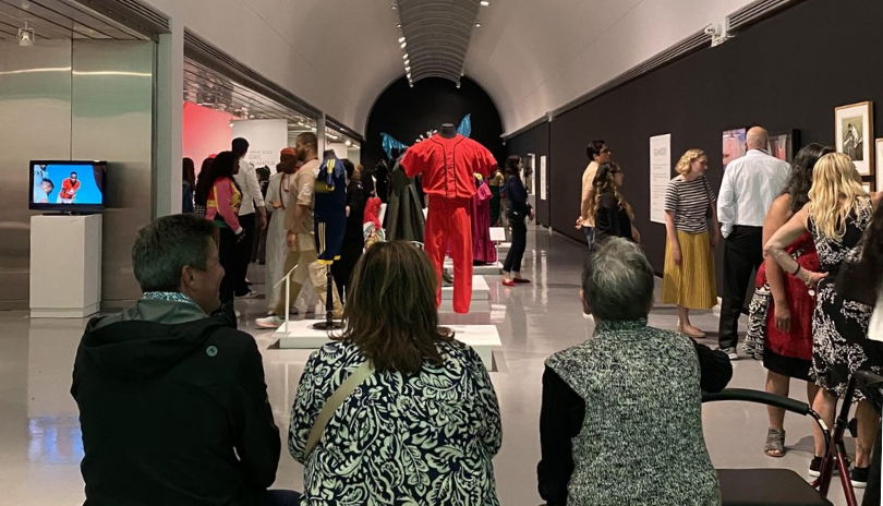Three people sit in a museum as others browse a fashion art exhibition.